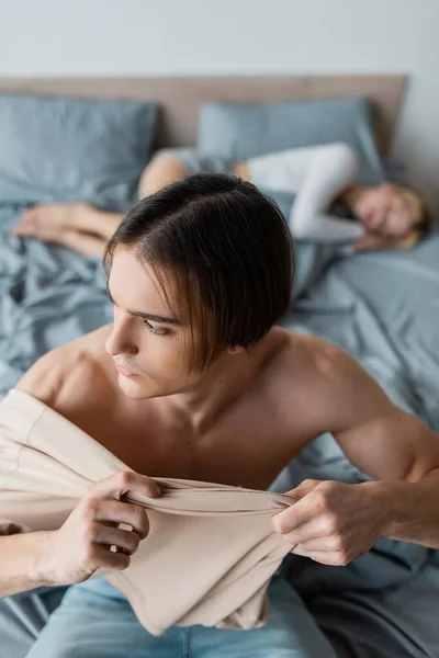 Мужчина без рубашки в одежде рядом с размытой женщиной, лежащей на кровати после одной ночи — стоковое фото