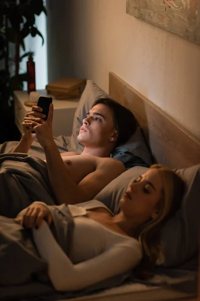 Homem infiel usando telefone celular perto de namorada loira deitada na cama, traindo conceito — Fotografia de Stock