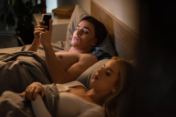 Unfaithful man using mobile phone and looking at blonde girlfriend sleeping in bed, cheating concept — Stock Photo