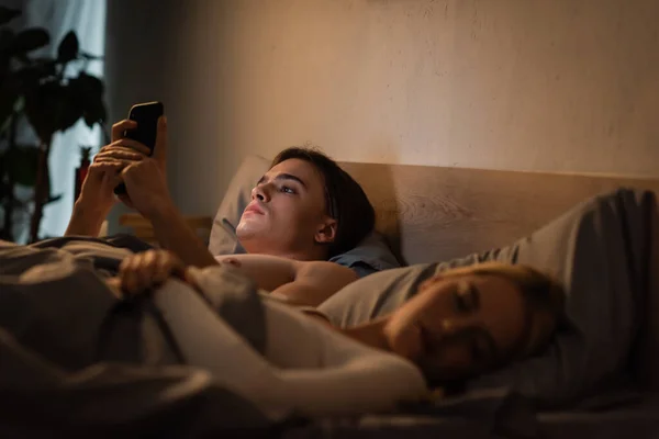 Jovem homem usando telefone celular ao lado de namorada loira dormindo na cama, traindo conceito — Fotografia de Stock