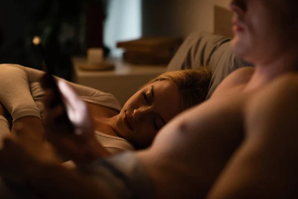 Mujer rubia durmiendo al lado de novio infiel usando smartphone, concepto de engaño - foto de stock