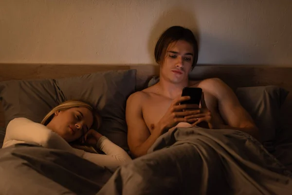 Awakened man using mobile phone next to blonde woman sleeping at night, cheating concept — Stock Photo