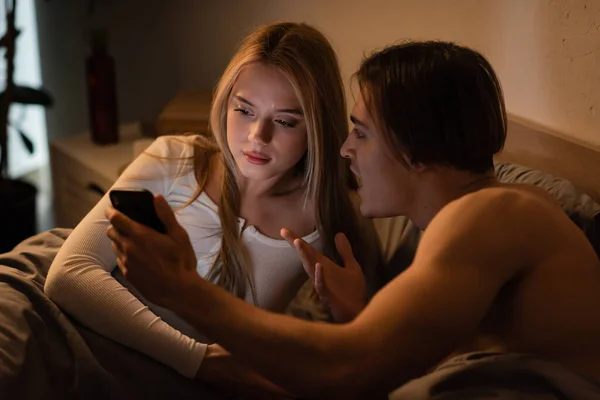 Uomo senza maglietta che tiene smartphone e litiga con la fidanzata infedele in camera da letto — Foto stock