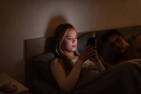 Mensajería mujer rubia en el teléfono inteligente al lado de novio dormido por la noche - foto de stock