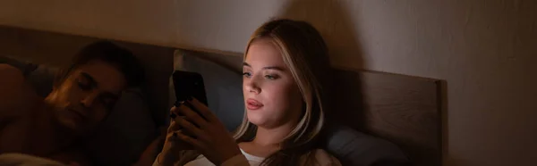 Blondine Nachrichten auf dem Smartphone neben schlafendem Freund in der Nacht, Banner — Stockfoto