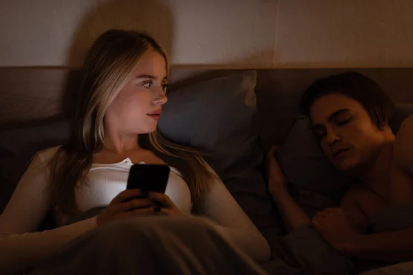Blonde woman using smartphone next to sleeping boyfriend at night, cheating concept — Stock Photo