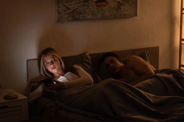 Mujer infiel mensajes de texto en el teléfono inteligente junto a dormir novio por la noche, concepto de engaño - foto de stock