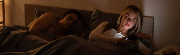 Donna bionda messaggistica su smartphone accanto al fidanzato addormentato di notte, banner — Foto stock