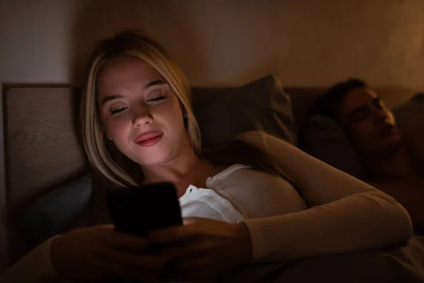 Fröhliche Frau, die nachts neben ihrem schlafenden Freund auf dem Smartphone Nachrichten sendet — Stockfoto