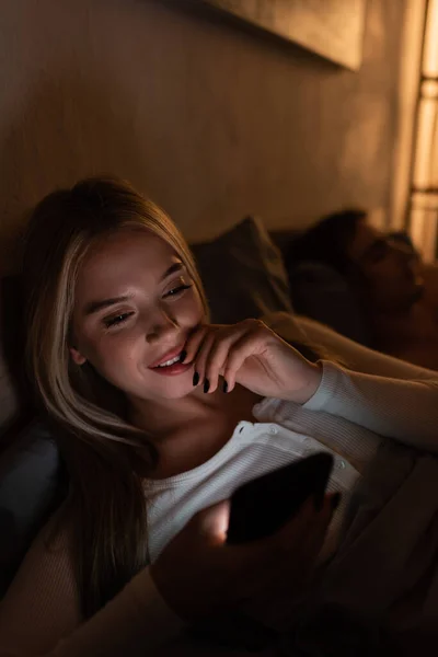 Усміхнена жінка обмінюється повідомленнями на смартфоні поруч зі сплячим хлопцем вночі — стокове фото