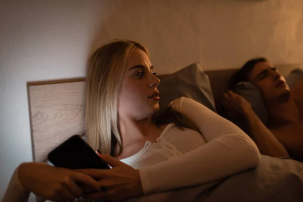 Mulher desleal usando smartphone ao lado de namorado adormecido à noite — Fotografia de Stock