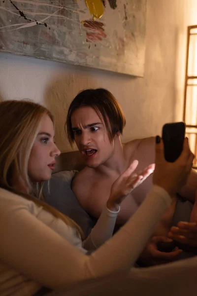Blondine hält Handy in der Hand, während sie mit wütendem Freund im Schlafzimmer streitet und betrügt Konzept — Stockfoto