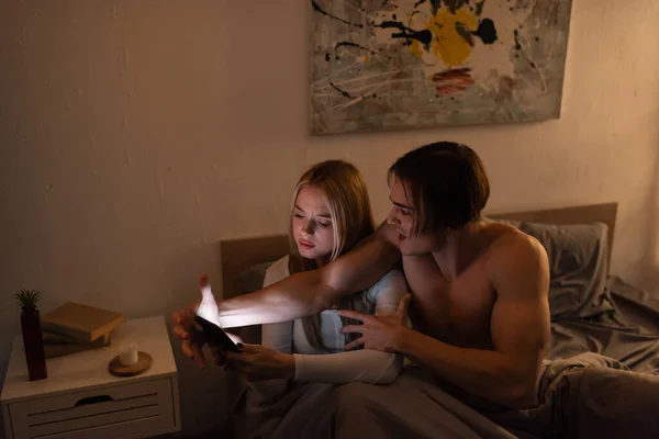 Wütender Mann schreit, während er mit blonder Freundin streitet, die Smartphone im Schlafzimmer hält, betrügt Konzept — Stockfoto