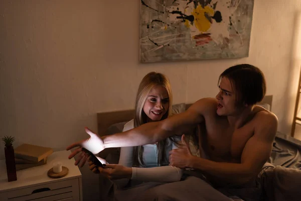 Hombre emocional peleando con novia preocupada sosteniendo teléfono inteligente en el dormitorio, concepto de engaño - foto de stock