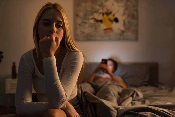 Worried woman sitting on bed near blurred boyfriend using smartphone in bedroom — Stock Photo