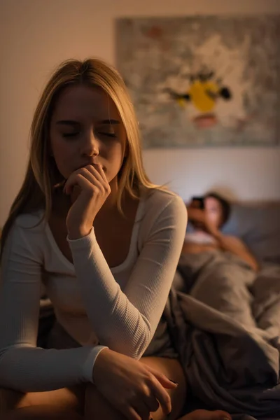 Worried woman sitting on bed near blurred boyfriend in bedroom, cheating concept — Stock Photo