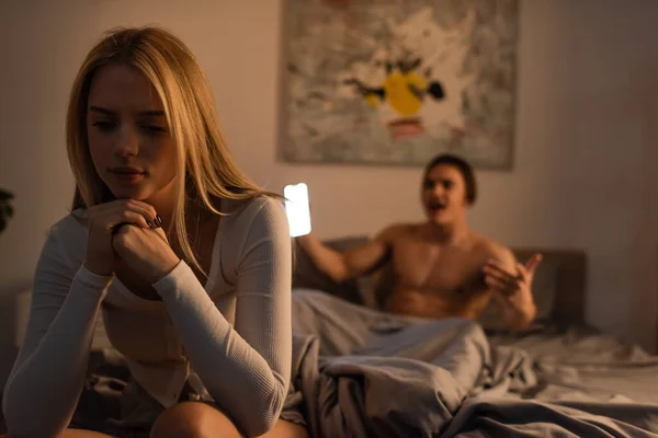 Mujer preocupada sentada en la cama cerca de novio gritando usando teléfono inteligente en el dormitorio, concepto de trampa - foto de stock