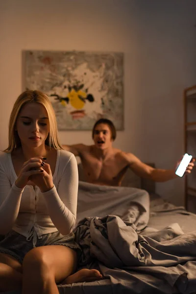 Блондинка сидит на кровати рядом с кричащим бойфрендом держа смартфон в спальне, обман концепции — стоковое фото