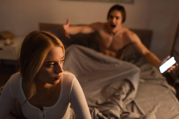 Mujer rubia sentada junto a novio emocional con smartphone en el dormitorio, concepto de engaño - foto de stock