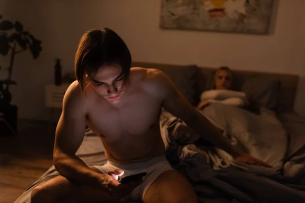 Uomo senza maglietta utilizzando il telefono cellulare mentre seduto sul letto vicino fidanzata su sfondo sfocato, concetto di tradimento — Foto stock