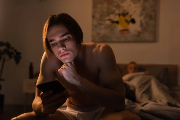 Uomo senza maglietta utilizzando il cellulare mentre seduto sul letto vicino fidanzata su sfondo sfocato, concetto di tradimento — Foto stock