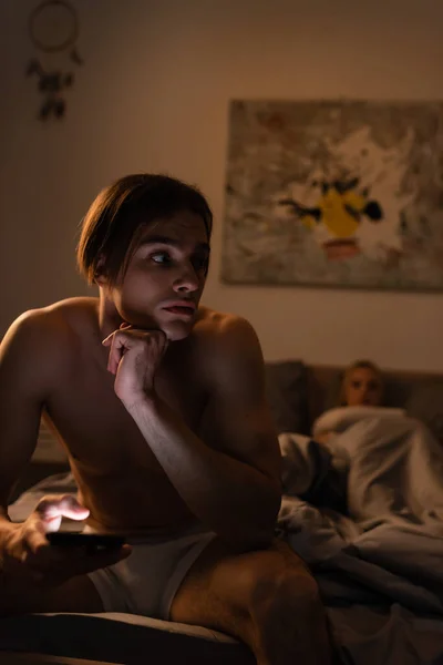 Hemdloser Mann benutzt Handy, während er neben Freundin auf verschwommenem Hintergrund sitzt, Schummel-Konzept — Stockfoto
