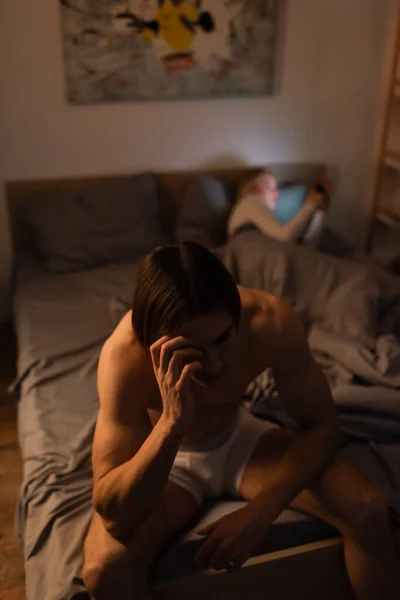 Sad and shirtless man sitting on bed near blurred girlfriend, cheating concept — Stock Photo