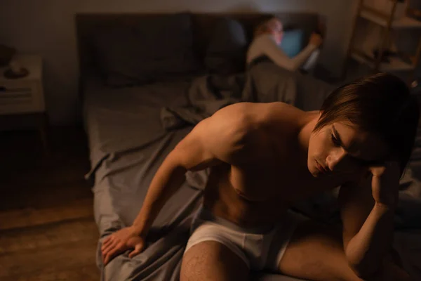 Aufgebrachter Mann in Unterwäsche sitzt auf Bett neben verschwommener Freundin und betrügt Konzept — Stockfoto
