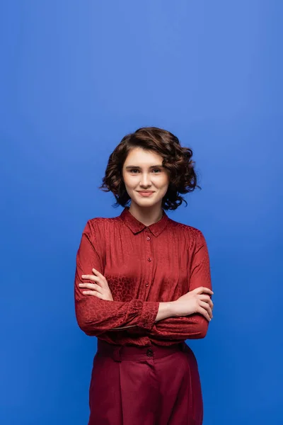 Cheerful language teacher with short curly hair standing with folded arms isolated on blue — Stock Photo