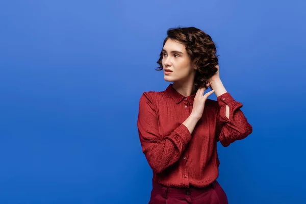 Thoughtful language teacher in red blouse adjusting curly hair and looking away isolated on blue — Stock Photo