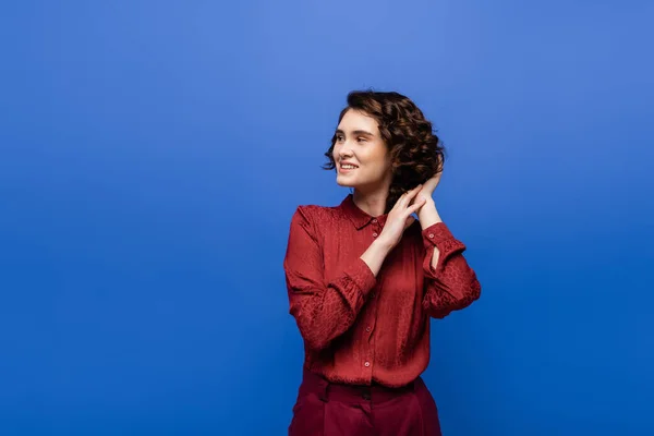 Pleased language teacher in red blouse adjusting curly hair and looking away isolated on blue — Stock Photo