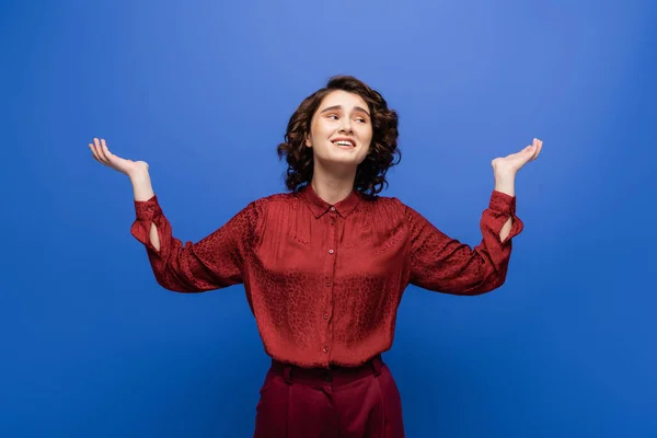 Smiling young woman with short curly hair gesturing while looking away isolated on blue — Stock Photo