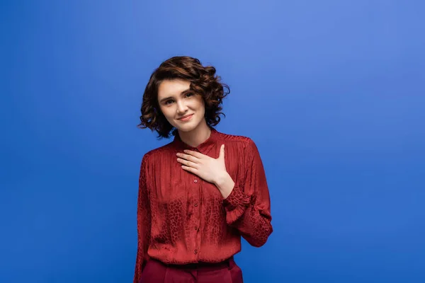 Thankful language teacher with short curly hair holding hand on chest isolated on blue — Stock Photo