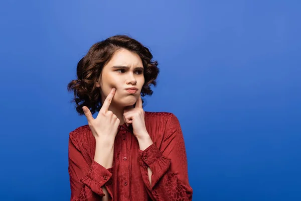 Unzufriedene junge Frau zeigt mit den Fingern auf aufgeblasene Wangen, während sie vereinzelt auf blauem Grund wegschaut — Stockfoto