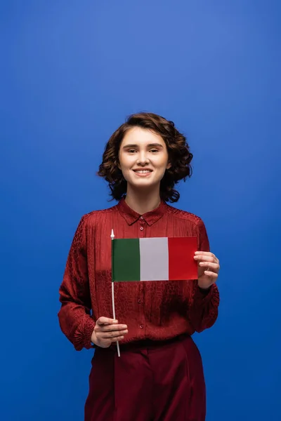 Happy language teacher smiling while holding flag of Italy and looking at camera isolated on blue — Stock Photo