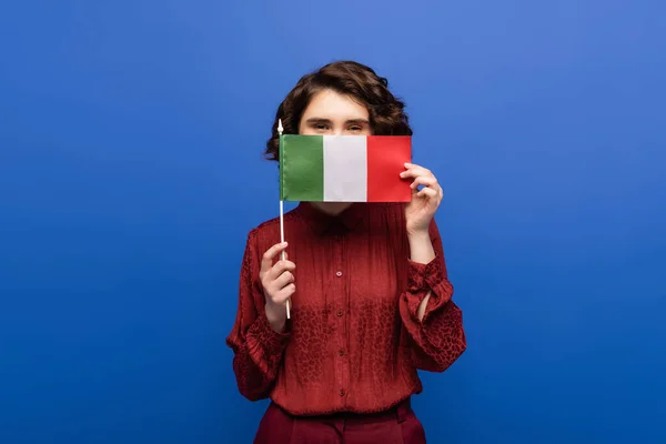 Professeur de langue bouclée couvrant le visage tout en tenant le drapeau de l'Italie et en regardant la caméra isolée sur bleu — Photo de stock
