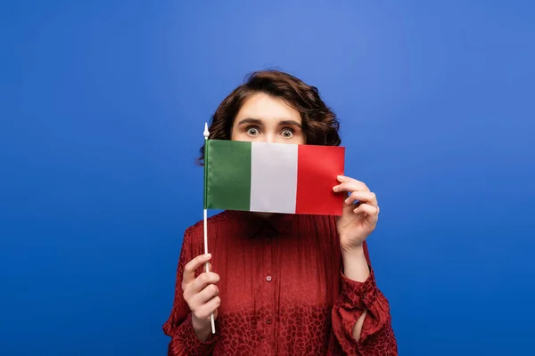 Jeune femme bouclée couvrant le visage tout en tenant le drapeau de l'Italie et en regardant la caméra isolée sur bleu — Photo de stock