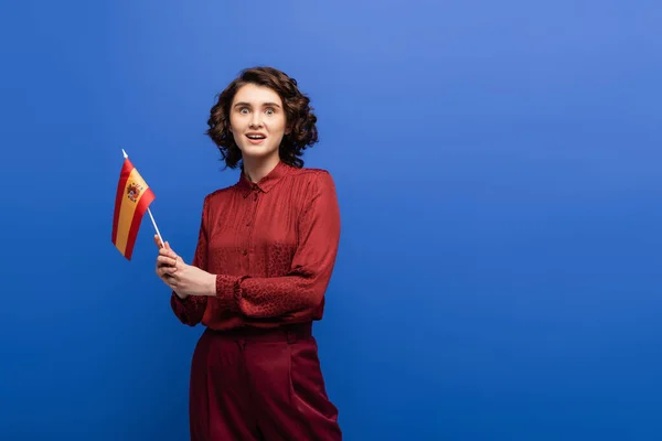Professeur de langue étonné regardant la caméra tout en tenant le drapeau de l'Espagne isolé sur bleu — Photo de stock