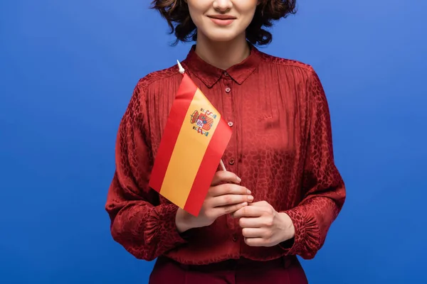 Vista recortada de la feliz profesora de idiomas sonriendo mientras sostiene la bandera de España aislada en azul - foto de stock