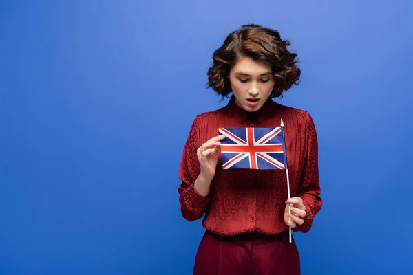 Shocked student with curly hair looking at flag of United Kingdom isolated on blue — Stock Photo