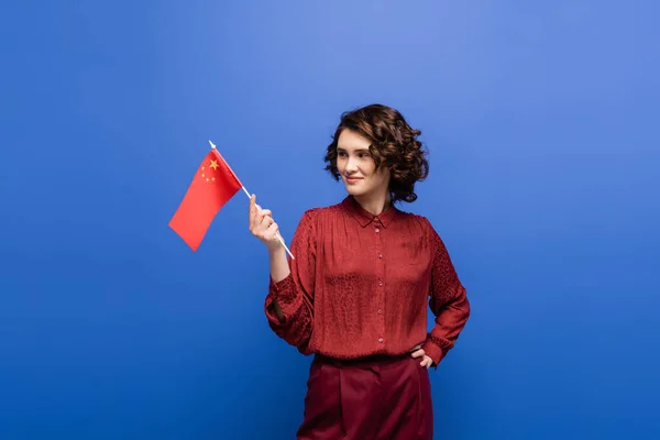 Professeur de langue heureux tenant drapeau de la Chine tout en se tenant la main sur la hanche isolé sur bleu — Photo de stock