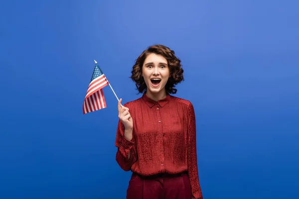 Взволнованная женщина с открытым ртом держа флаг США и глядя на камеру изолированы на синий — стоковое фото