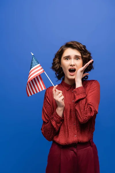 Шокований студент з відкритим ротом тримає прапор США і дивиться на камеру ізольовано на синьому — стокове фото