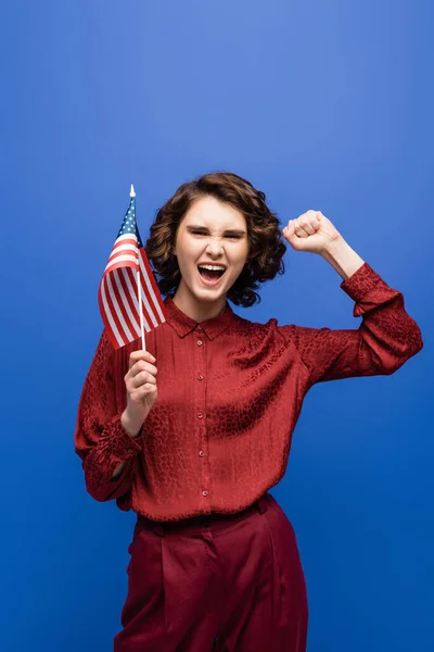 Estudiante emocionado mostrando el gesto de éxito mientras sostiene la bandera de EE.UU. y mirando a la cámara aislada en azul - foto de stock