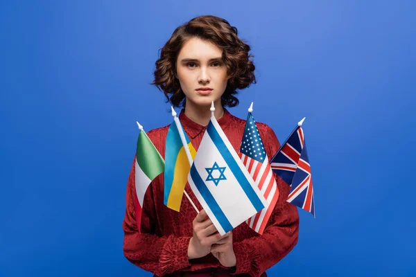 Young and confident woman looking at camera while holding international flags isolated on blue — Stock Photo