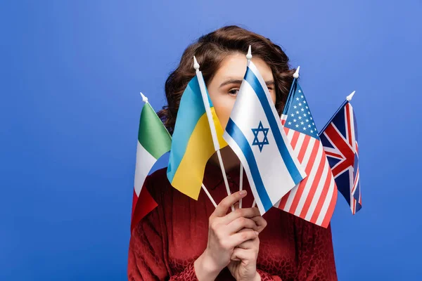 Jeune femme regardant caméra derrière divers drapeaux internationaux isolés sur bleu — Photo de stock