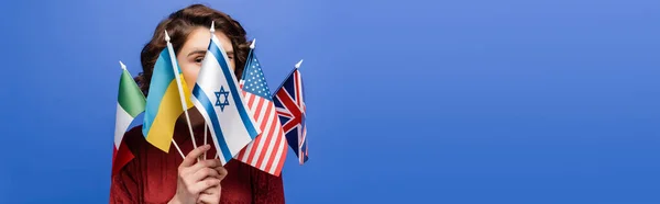 Jeune femme obscurcissant le visage avec différents drapeaux et regardant la caméra isolée sur bleu, bannière — Photo de stock