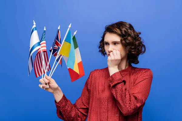 Nachdenkliche Frau, die die Hand vor dem Gesicht hält, während sie Flaggen verschiedener Länder betrachtet, die auf blauem Grund isoliert sind — Stockfoto