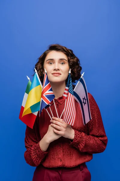 Mujer asombrada sosteniendo banderas de varios países y mirando a la cámara aislada en azul - foto de stock