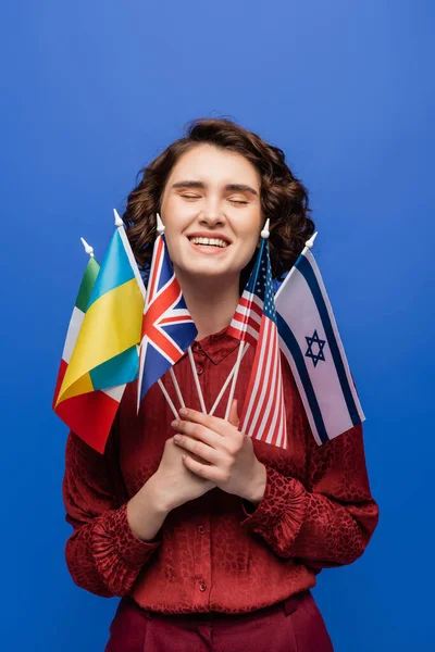 Happy young woman holding international flags and smiling with closed eyes isolated on blue — Stock Photo